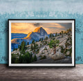 A framed photograph depicting a mountainous landscape with a large, snow-capped peak in the distance, surrounded by trees and rocky terrain. The sky is filled with warm, golden light, creating a serene and picturesque scene.