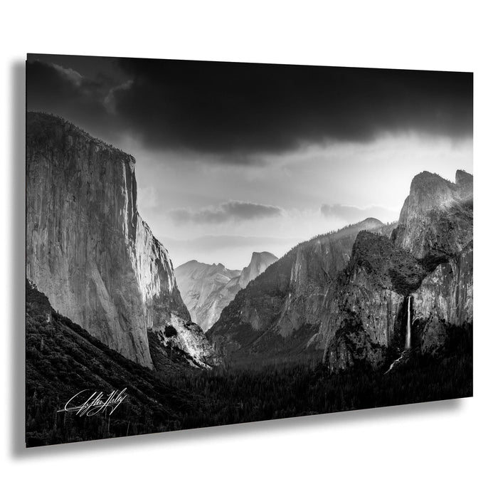 A dramatic black and white landscape photograph depicting a mountainous, forested valley with towering cliffs and a waterfall cascading down the side.
