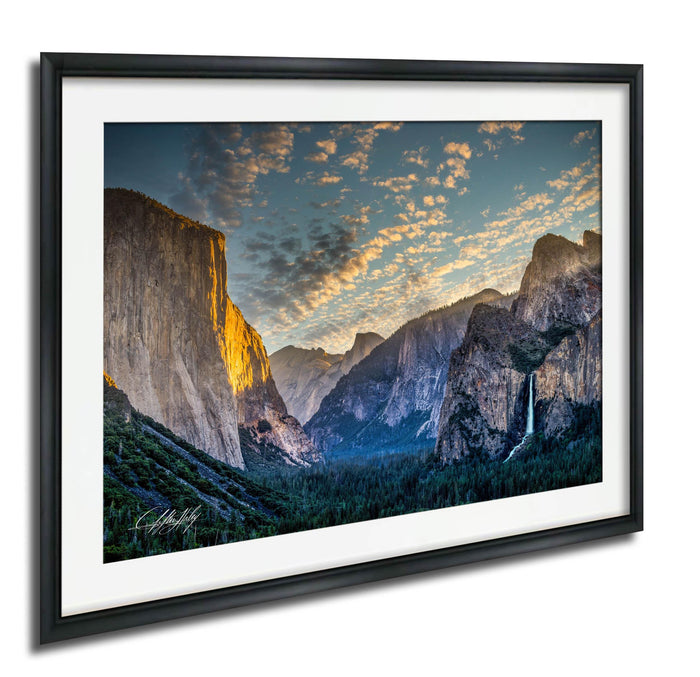 a breathtaking view of the majestic Yosemite Falls, with its cascading waters flowing down the towering granite cliffs. The sky above is a beautiful canvas of blue and orange hues, creating a stunning backdrop for the natural landscape. The photograph is framed and hangs on a wall, capturing the essence of this awe-inspiring scene.