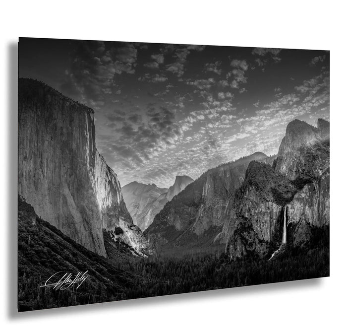 A dramatic black and white landscape photograph depicting a mountainous, forested valley with towering cliffs and a waterfall cascading down the side of one of the cliffs.